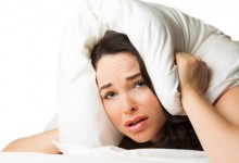 A tired sleepless woman puts the pillow over her head and looks helplessy at camera. Isolated over white.